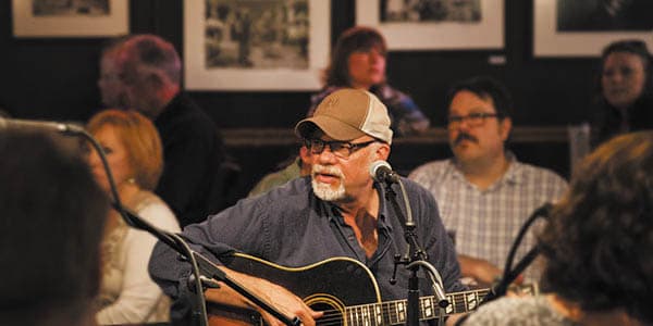 A man in black-rimmed glasses picks a guitar as an audience enjoys his music.