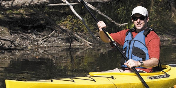 A man in a yellow kayak wearing a blue life vest and shades smiles at the camera.