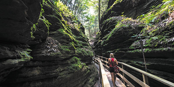 A woman walks along a wooden trail in a narrow ravine with moss-covered rock.