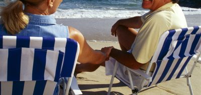 Blonde haired woman and tan older man holding hands sitting on beach chairs