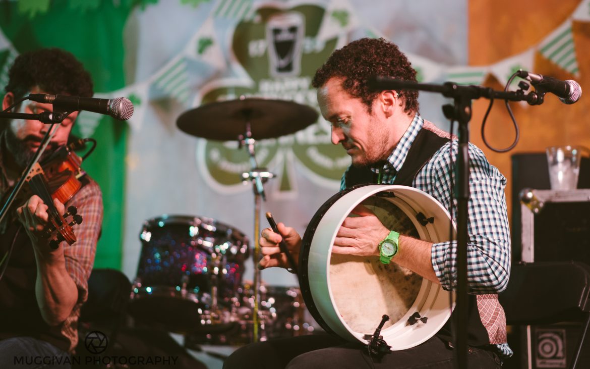Man playing large handheld drum on stage, with drum set in background
