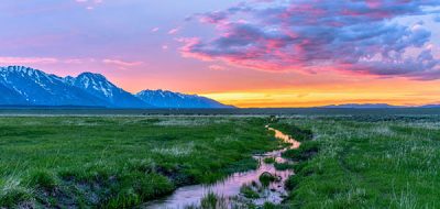 Colorful spring sunset at a green mountain field with a winding stream near Mormon Row historic district in Grand Teton National Park, Wyoming, USA.