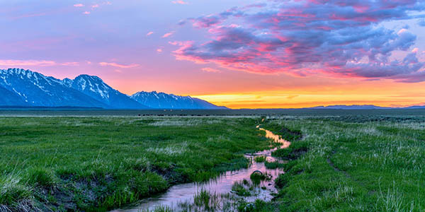 Colorful spring sunset at a green mountain field with a winding stream near Mormon Row historic district in Grand Teton National Park, Wyoming, USA.