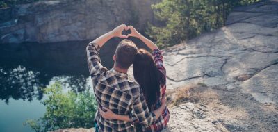 Good shiny morning in wood! Rear back high angle view of two people in love, wearing casual, sitting on the top of canyon stone rock, embracing, making heart shape with hands