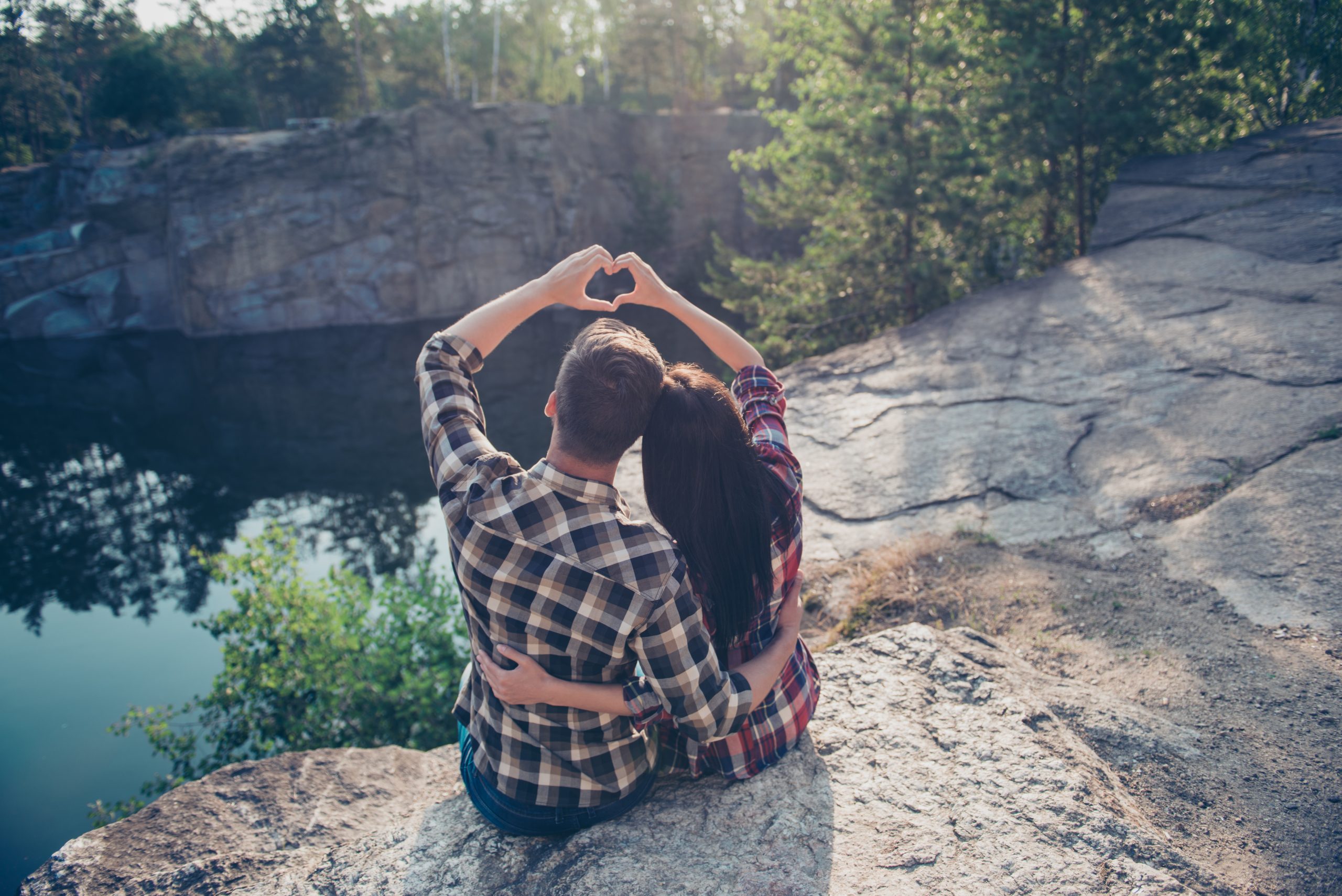 Good shiny morning in wood! Rear back high angle view of two people in love, wearing casual, sitting on the top of canyon stone rock, embracing, making heart shape with hands