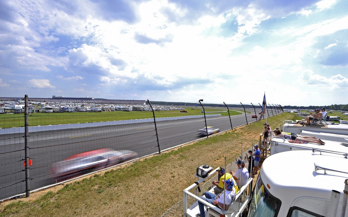 Stock cars zoom past a row of RVs with spectators perched on the roofs.