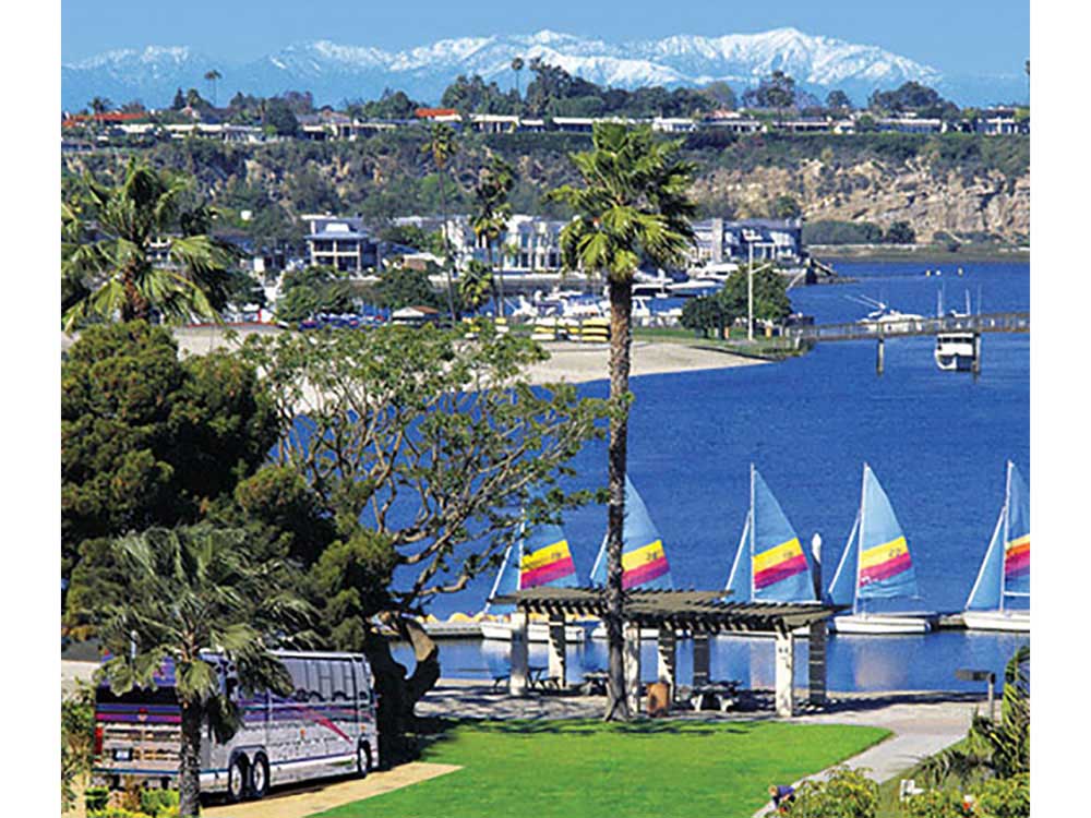 Boats with colorful triangular sails docked in Newport Bay with mountains rising in the background.