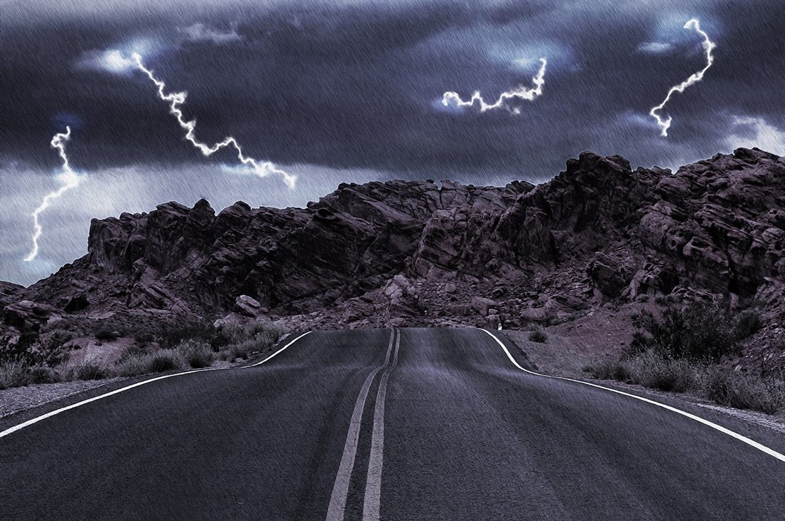 A two-lane road leads to a rocky horizon under flashing thunder.