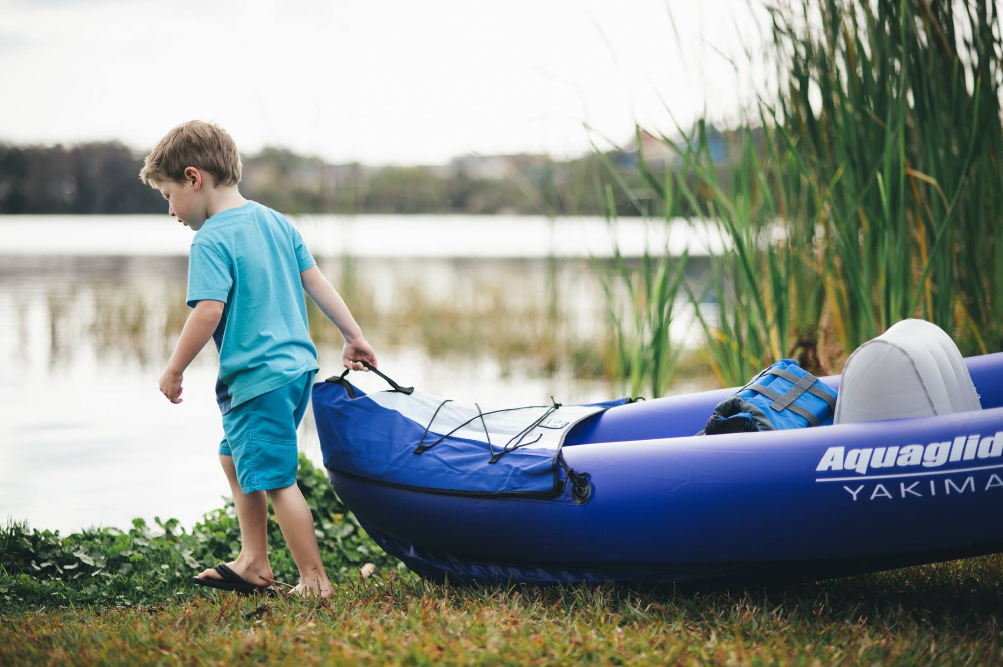 A boy drags an Aquaglide kayak across the grass toward a pond with reeds.