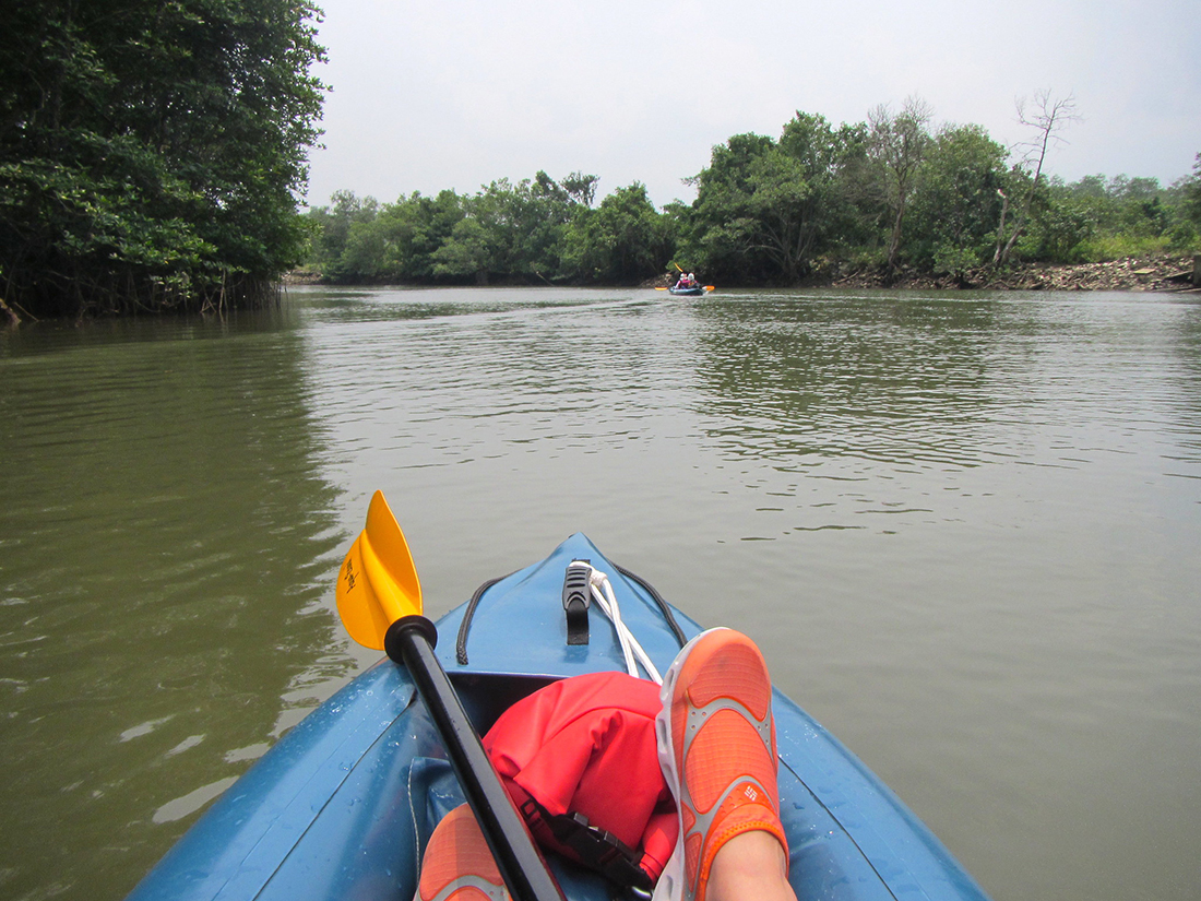 POV from kayak paddler: feet outstretched on muddy stream with paddles placed in resting position.