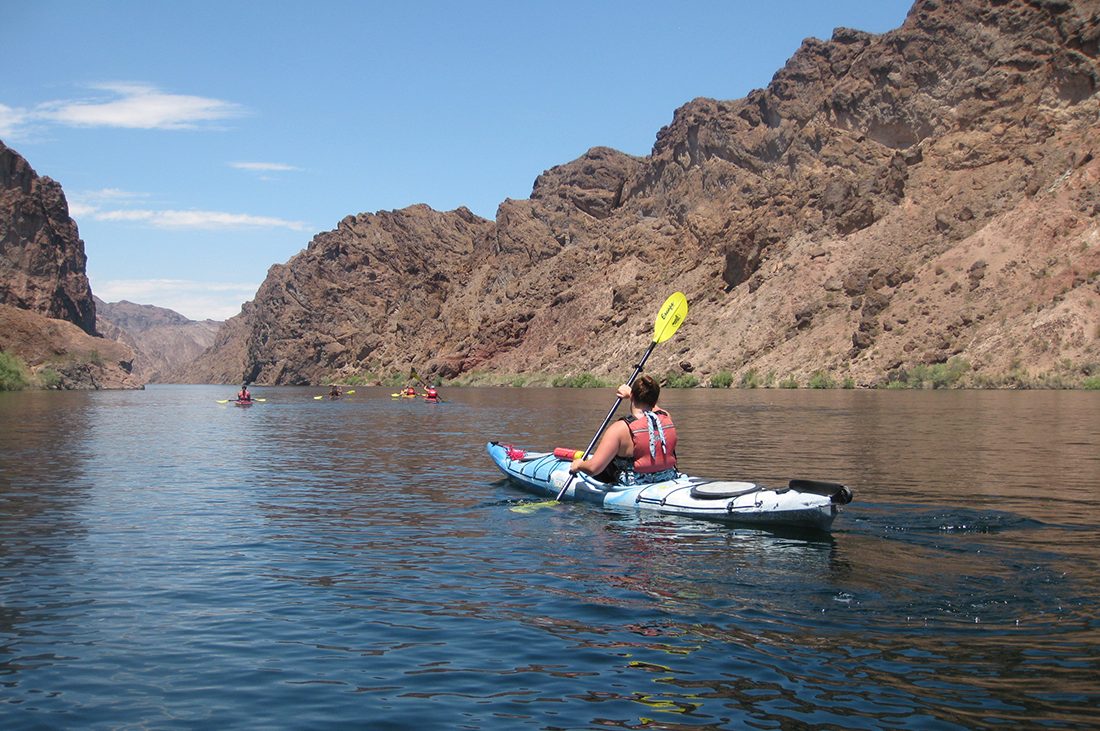 A kayaker knifes through the Colorado River with rocky banks rising on each side.