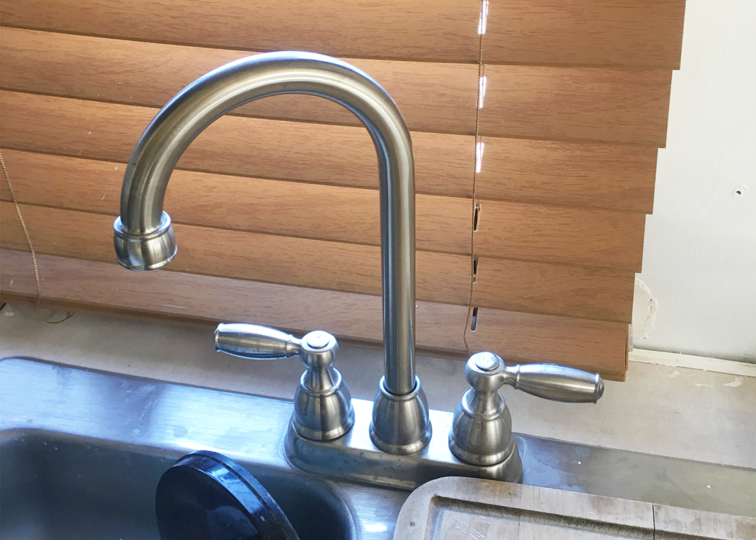 A kitchen faucet with wooden blinds in the background.