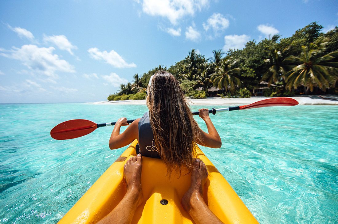 A girl sits in the front of a two-person kayak navigating clear waters in a tropical setting.