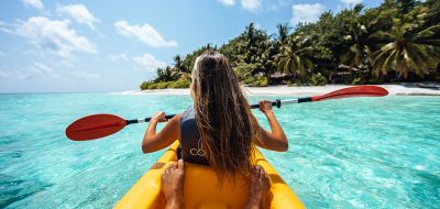 A girl sits in the front of a two-person kayak navigating clear waters in a tropical setting.