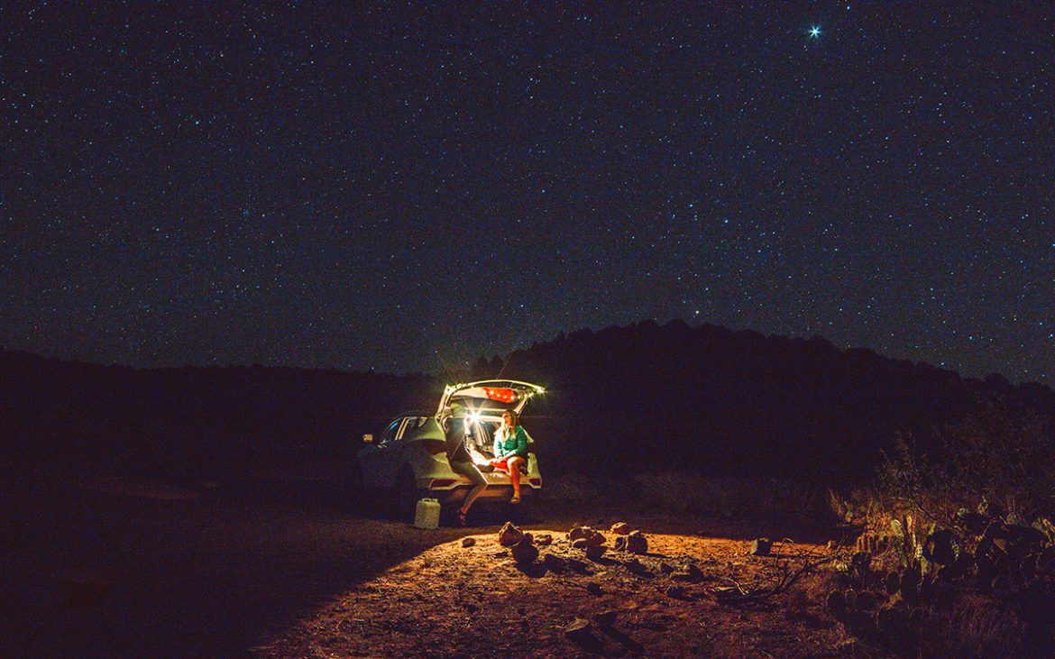 Two young women sit in the open hatchback of their vehicle in a desert campsite under a starry night sky.