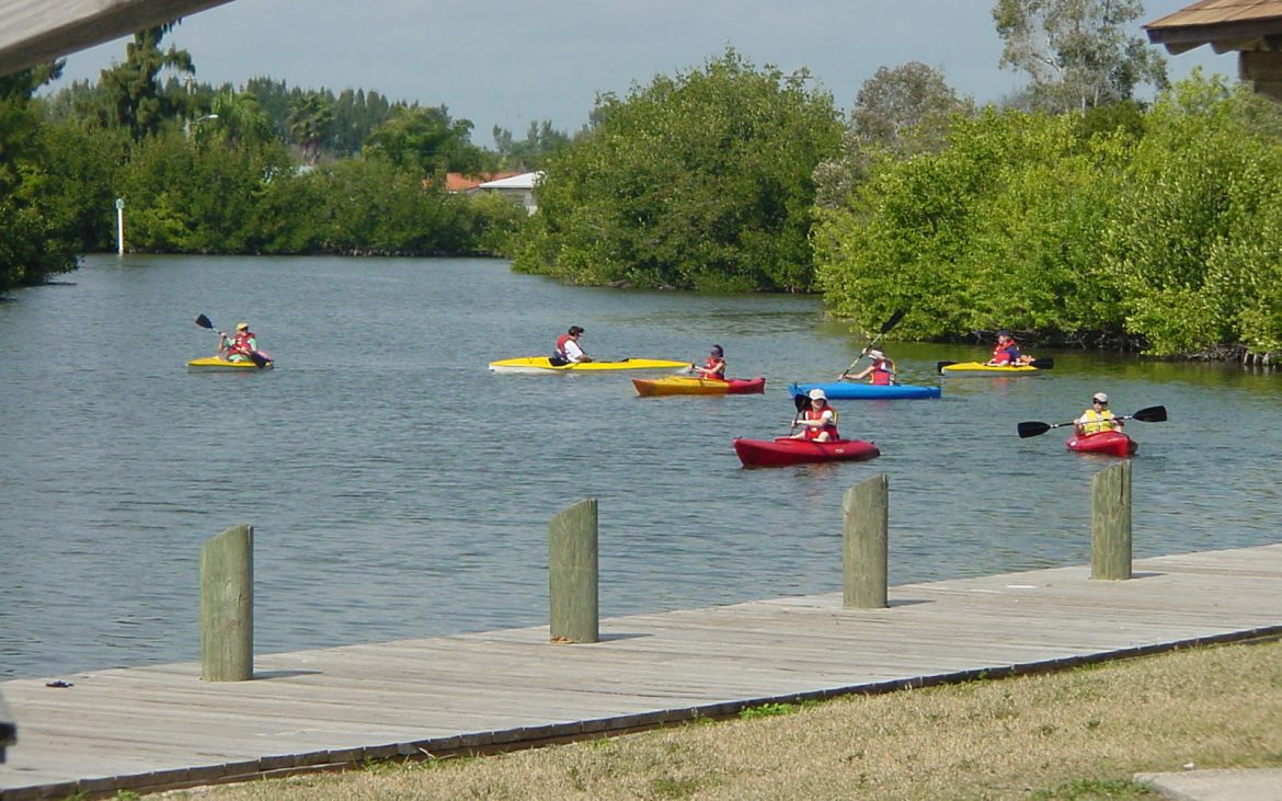 A group of kayakers navigate the bend of a river with dock in foreground.