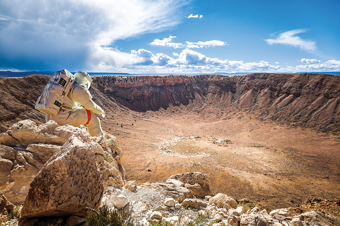 A person in a space suit looks out over Meteor Crater from the rim.