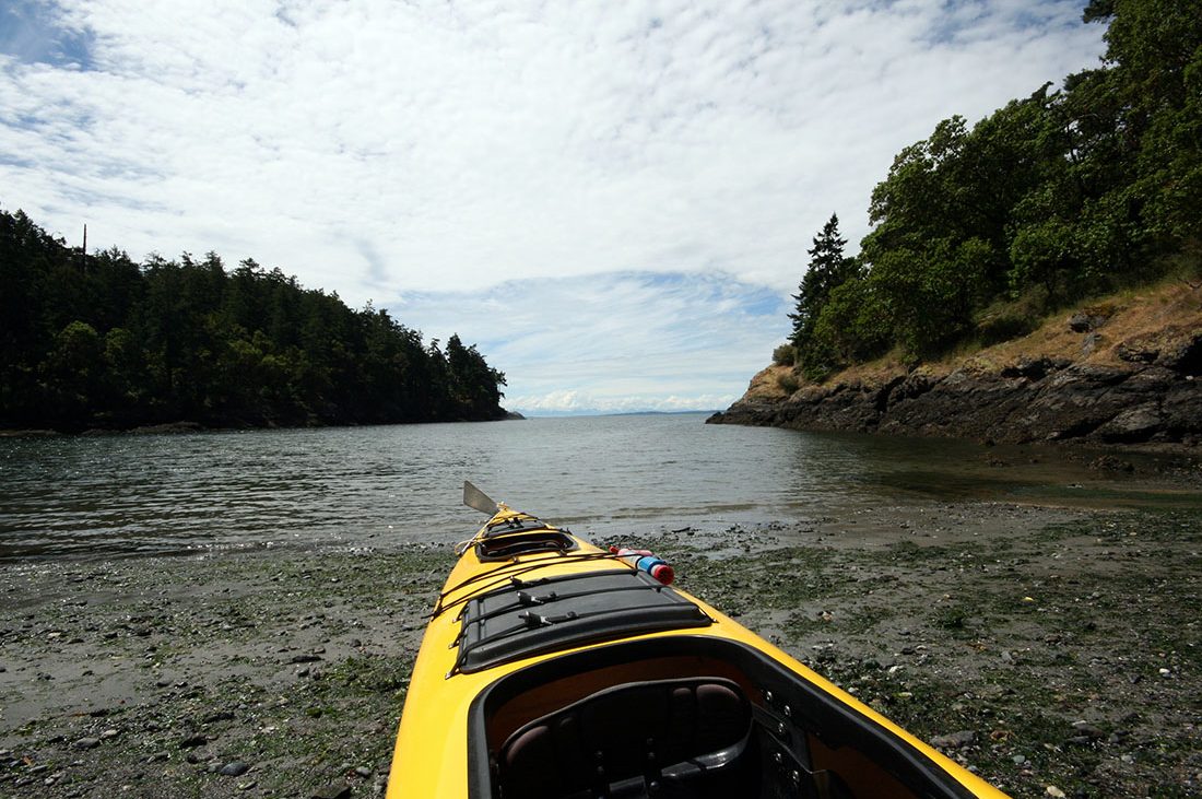 A kayak rests on the shore of an inlet surrounded by high banks and trees in the San Juan Islands.