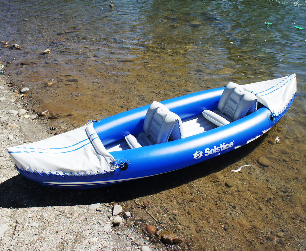 A beached blue Solstice two-seater kayak rests on the sandy shore of a clear waterway.
