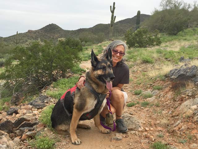 A woman on a desert trail poses with a hefty German Shepherd.