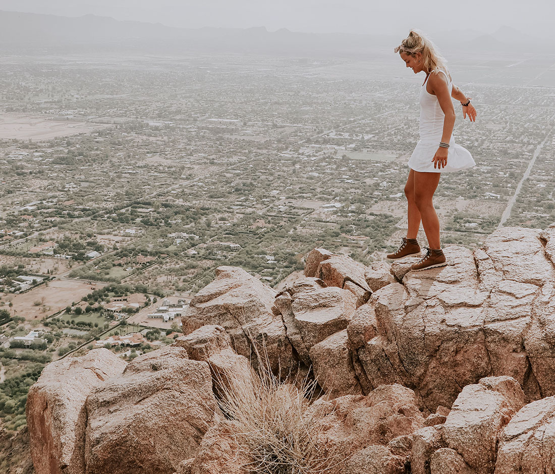 A woman in a white dress navigates the rocks on a mountaintop overlooking a city below.