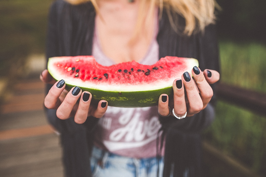 A woman with black nail polish holds up a slice of watermelon with black seeds. 