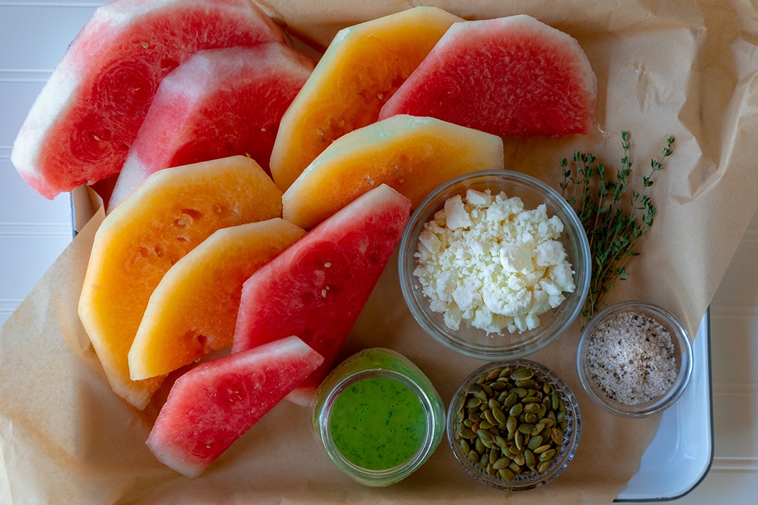 Slices of red and orange melon with small bowls of goat cheese, pepitas and more