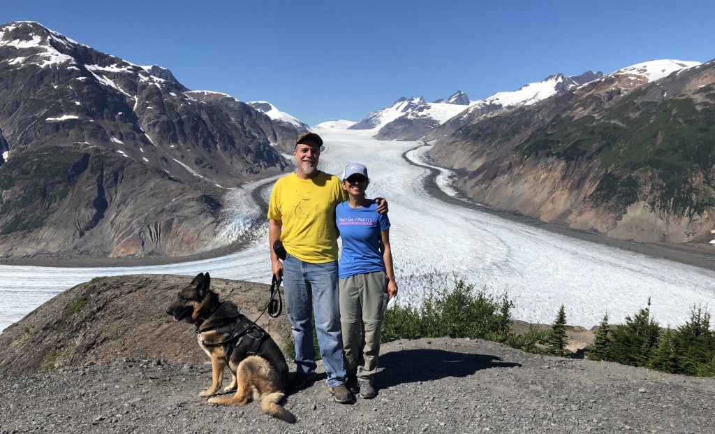 A couple stand on a ridge with their dog with a glacier in the background.