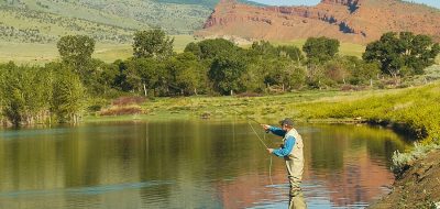 An angler with beige waders and a fishing vests casts a line in a lake as hills gently rise in the horizon.