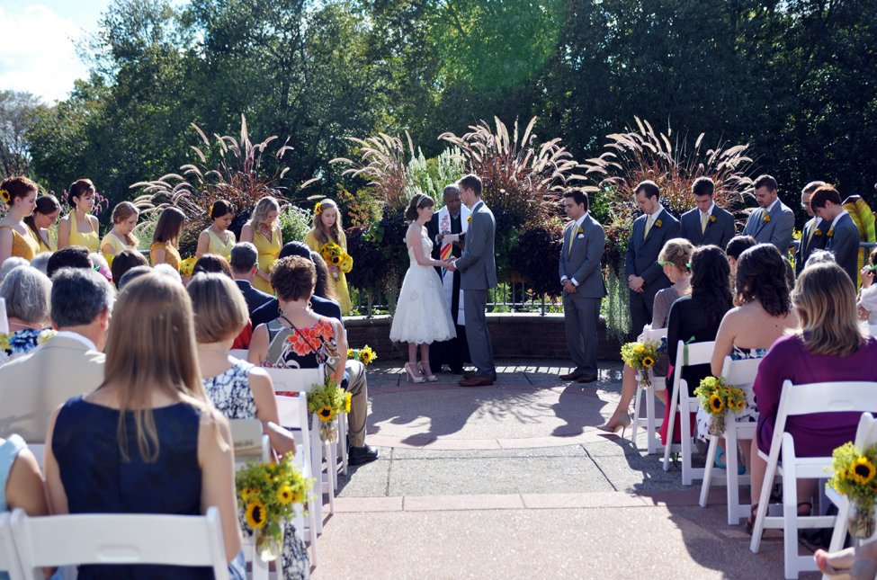 A couple getting married outdoors as guests look on amid plants and trees.