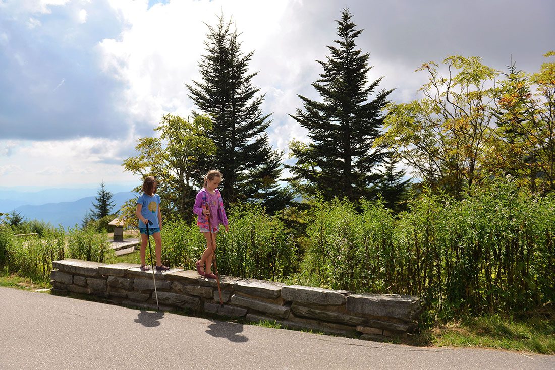 Two girls walk along a low brick wall with mountains and forests in the background.