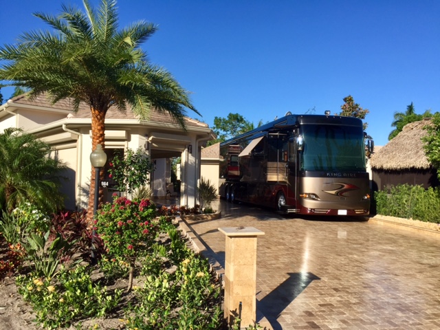 A black and tan motorhome parked in the tan brick driveway of a luxury house with palm tree in background.