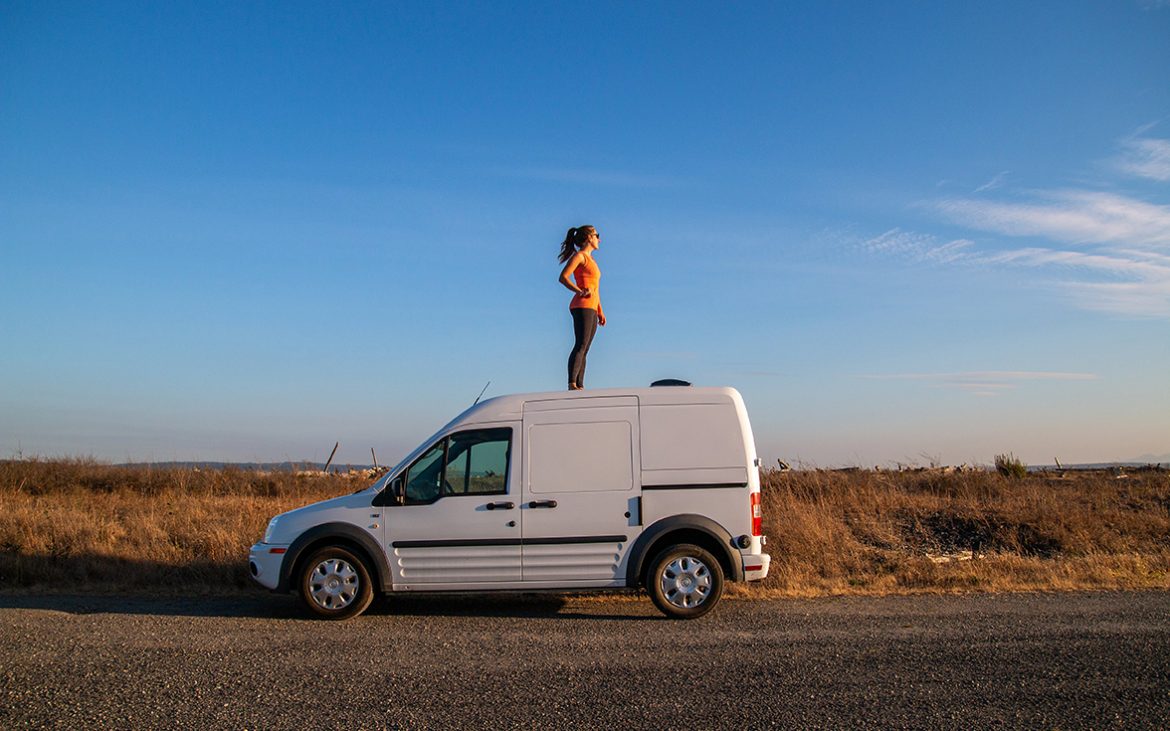 A solitary travelers stands on top of a camper van facing the sunset.