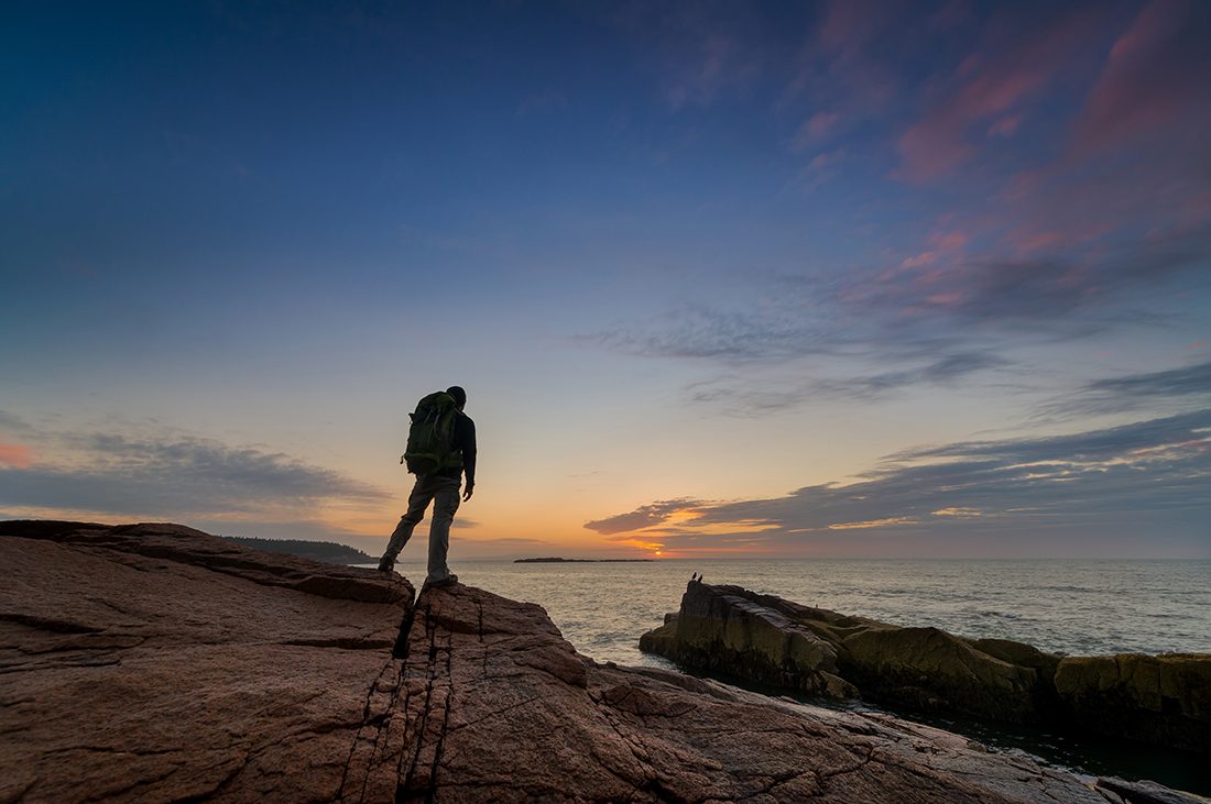 A man stands on rock outdropping facing the sun rising over the sea in Maine