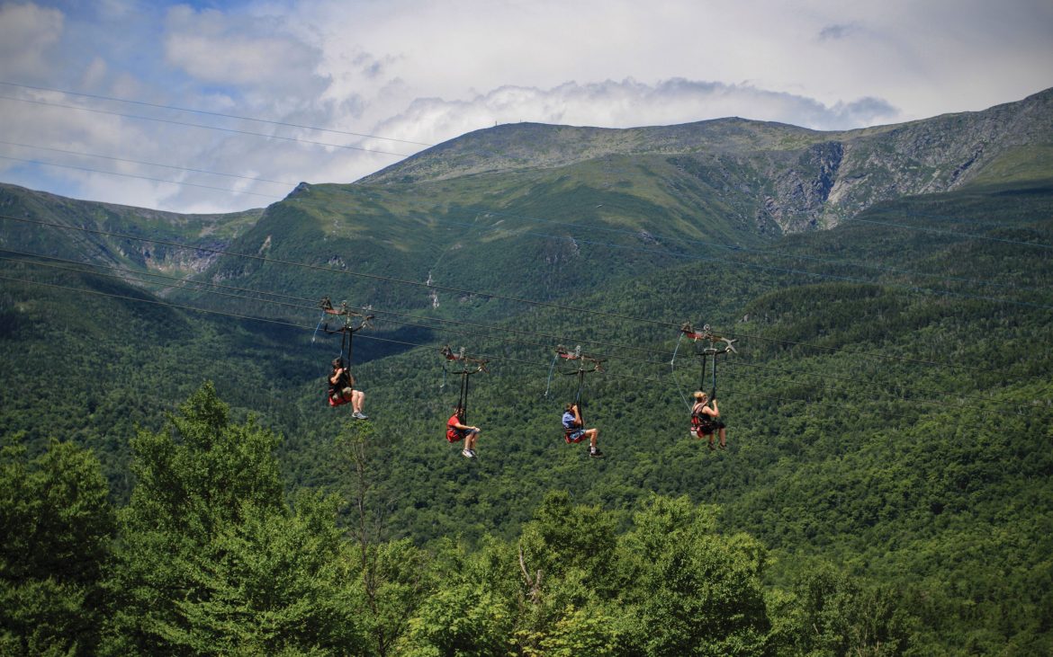 A group fo zipline riders careen over a green valley with mountains in the background.