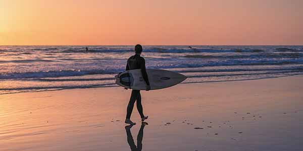 A surfer walks along the shore of Ocean Beach during sunset.