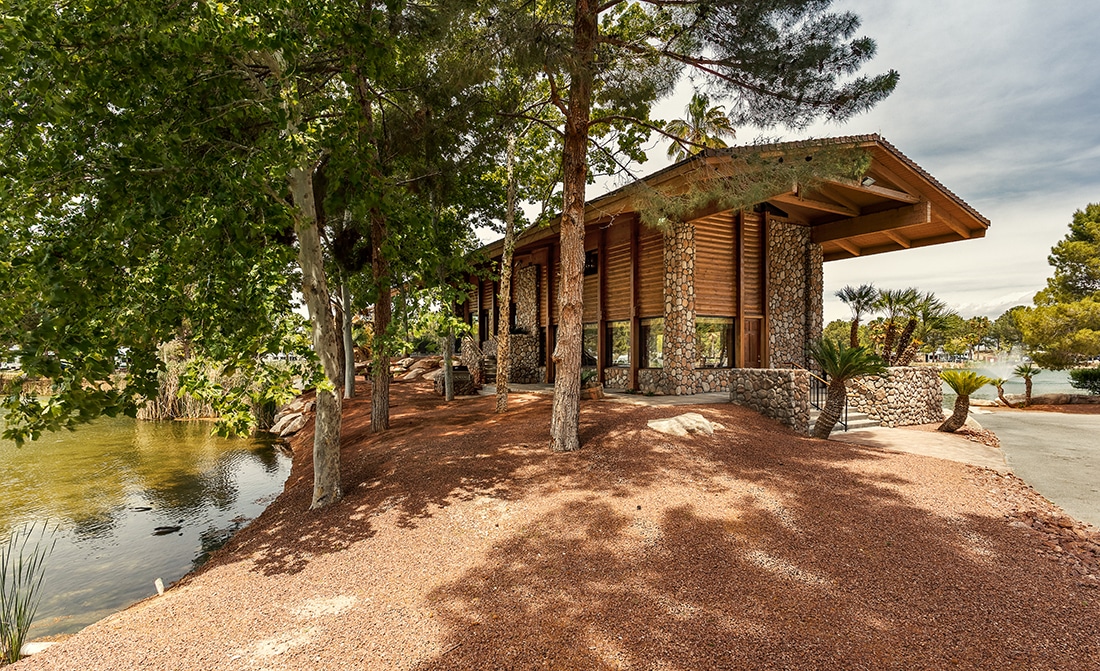 A thin stone and wood structure in the shade of all pine trees sits on the banks of a lake.