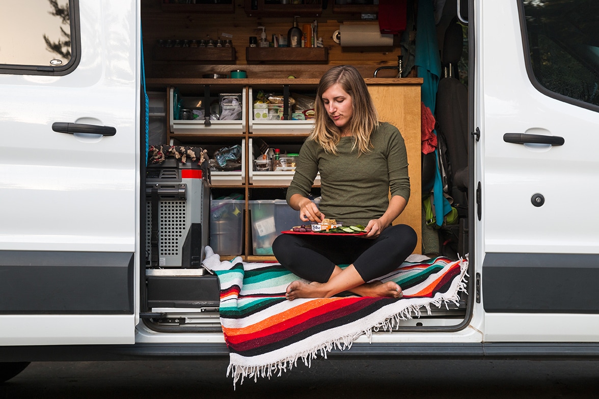A woman sits in the open door of her camper van on a Navajo blanket eating crackers and hummus.