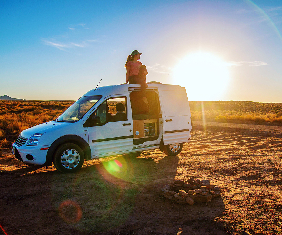 A woman wearing a cap sits on the roof of a camper van as the sun sets in the background.