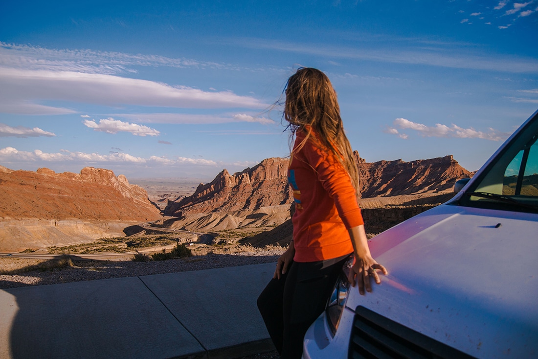 Minimalism on the road — a woman leans no the hood of a camper van and looks out on a rugged desert landscape.