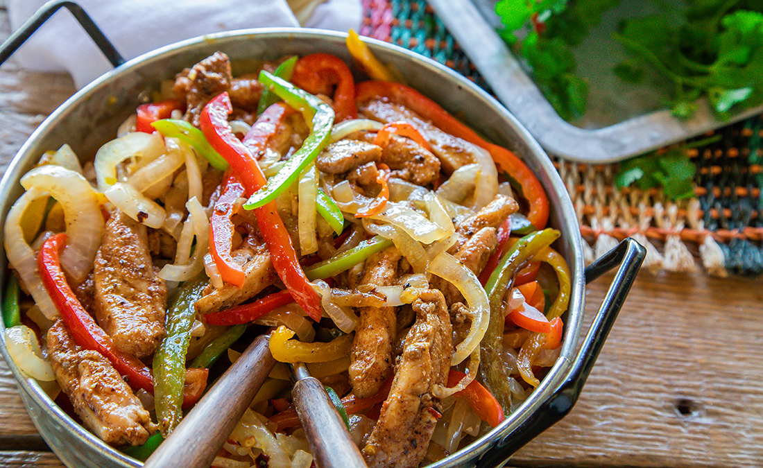 A bowl filled with cooked chicken, onions and peppers with serving utensils sticking out.