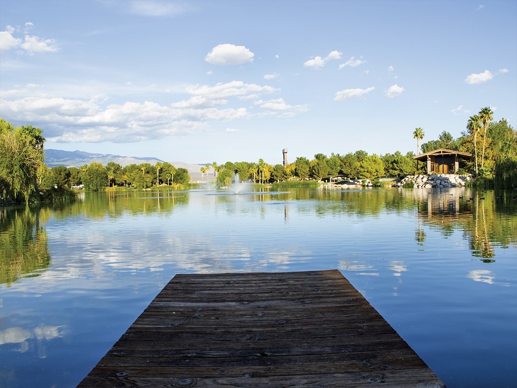 A wooden dock juts into a lake that reflects clear blue skies along with the palm trees that grow on the bank.