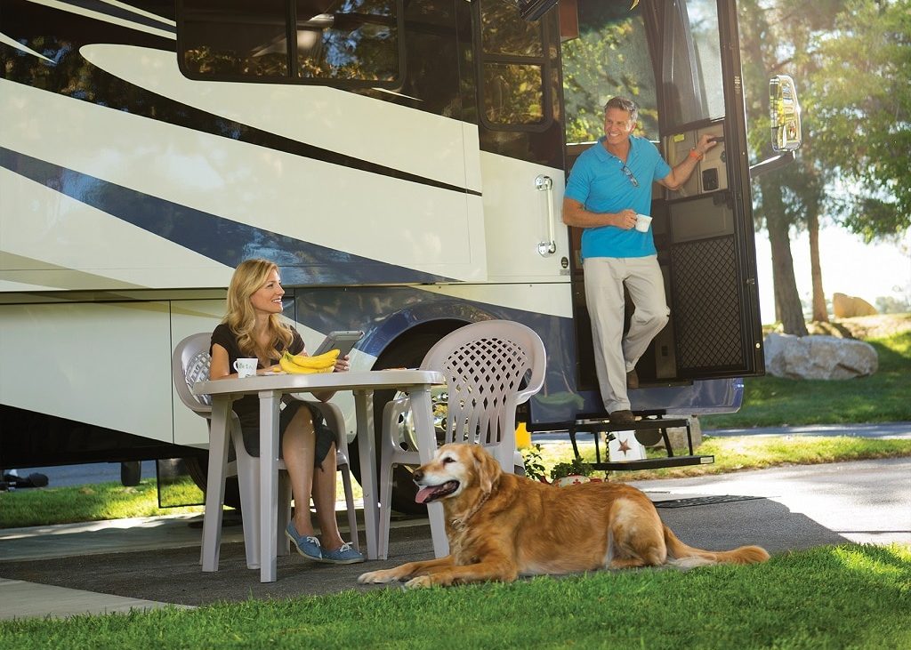 A couple lounge outside of their RV with a big dog sitting on the grass