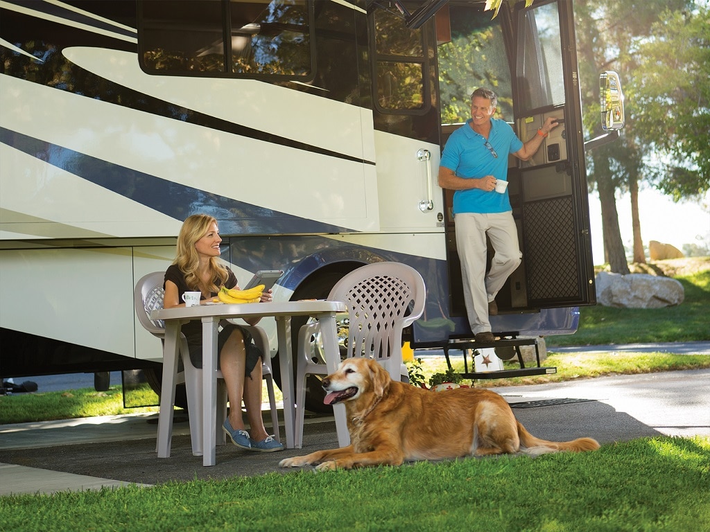 A couple lounge outside of their RV with a big dog sitting on the grass