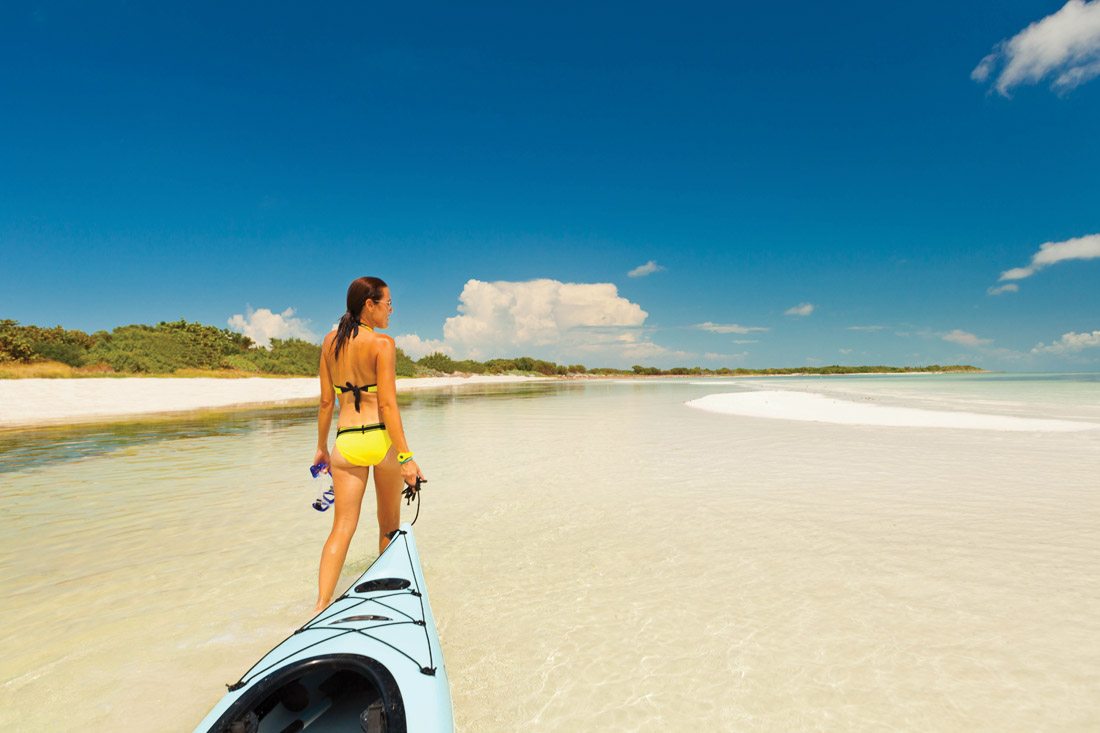 Woman in yellow bathing suit pulls her kayak along the shallows over clear water.