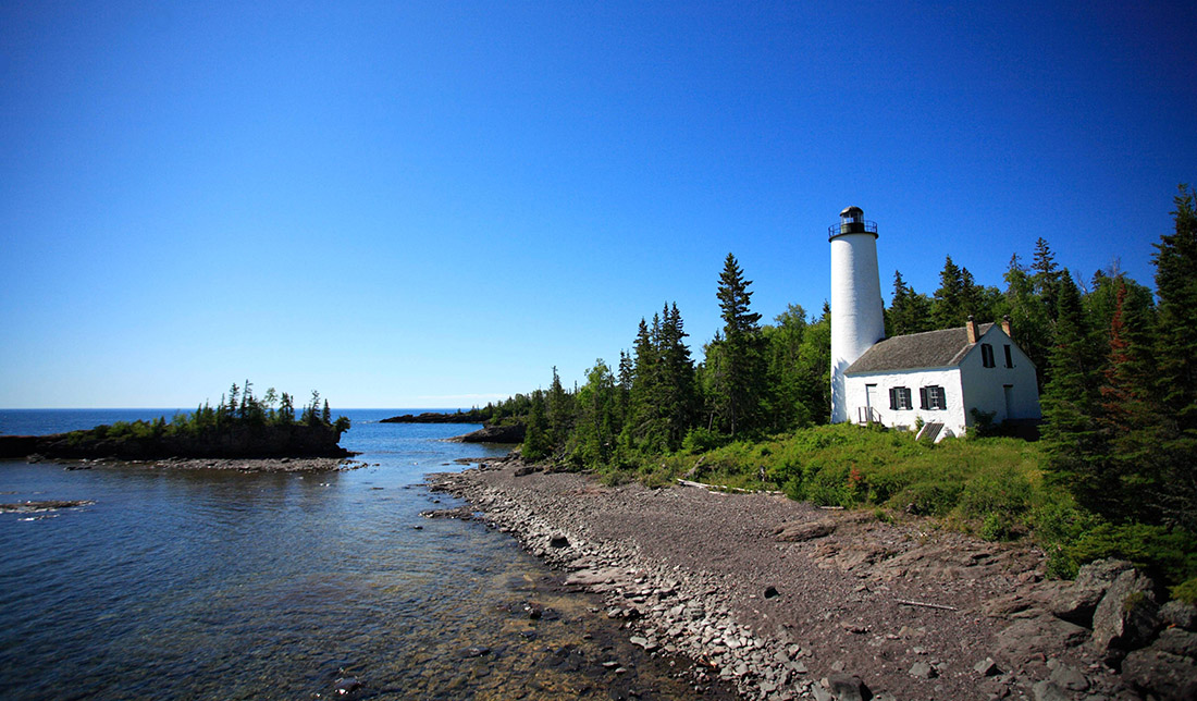 A lighthouse overlooks a rocky shoreline with forest in the background.