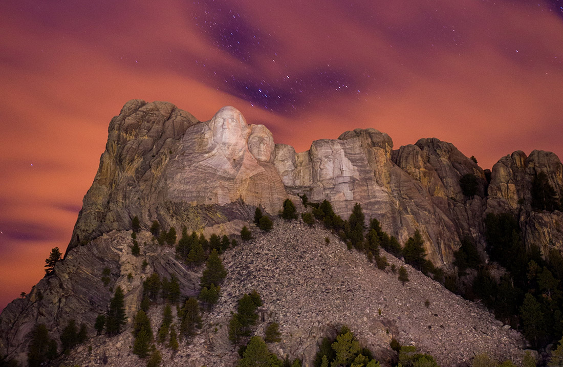 Four stone faces loom under a hazy night sky with gaps that reveal stars.