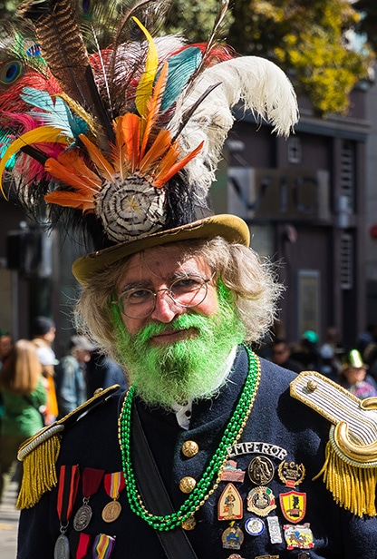 A dude with a green beard and feathered hat marches in an Irish parade.