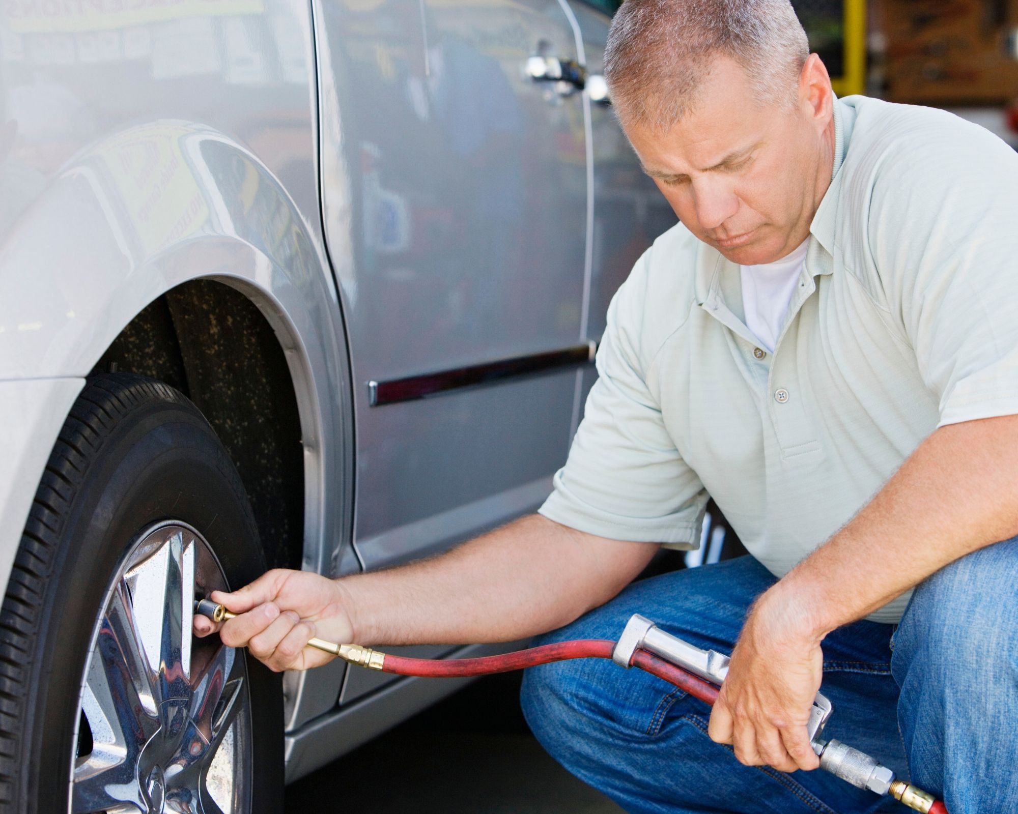 A squatting man pumps up the tire to a silver vehicle.