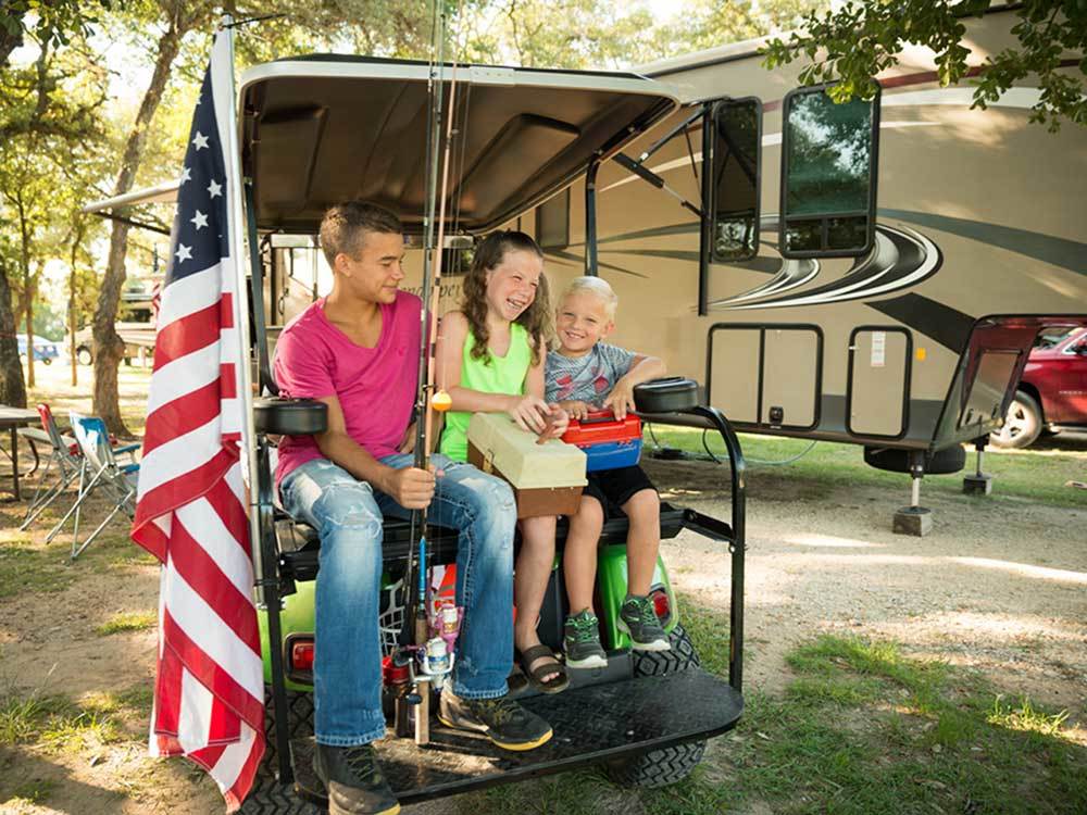 Three kids holding fishing poles sits in the back of an ATV with fifth-wheel in the background and American flag in the foreground.